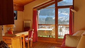 A cozy room with a wooden table, chairs, and a sofa. A large window offers a view of a snowy mountain landscape. - Apartment in the Megeve valley (Praz-sur-Arly)