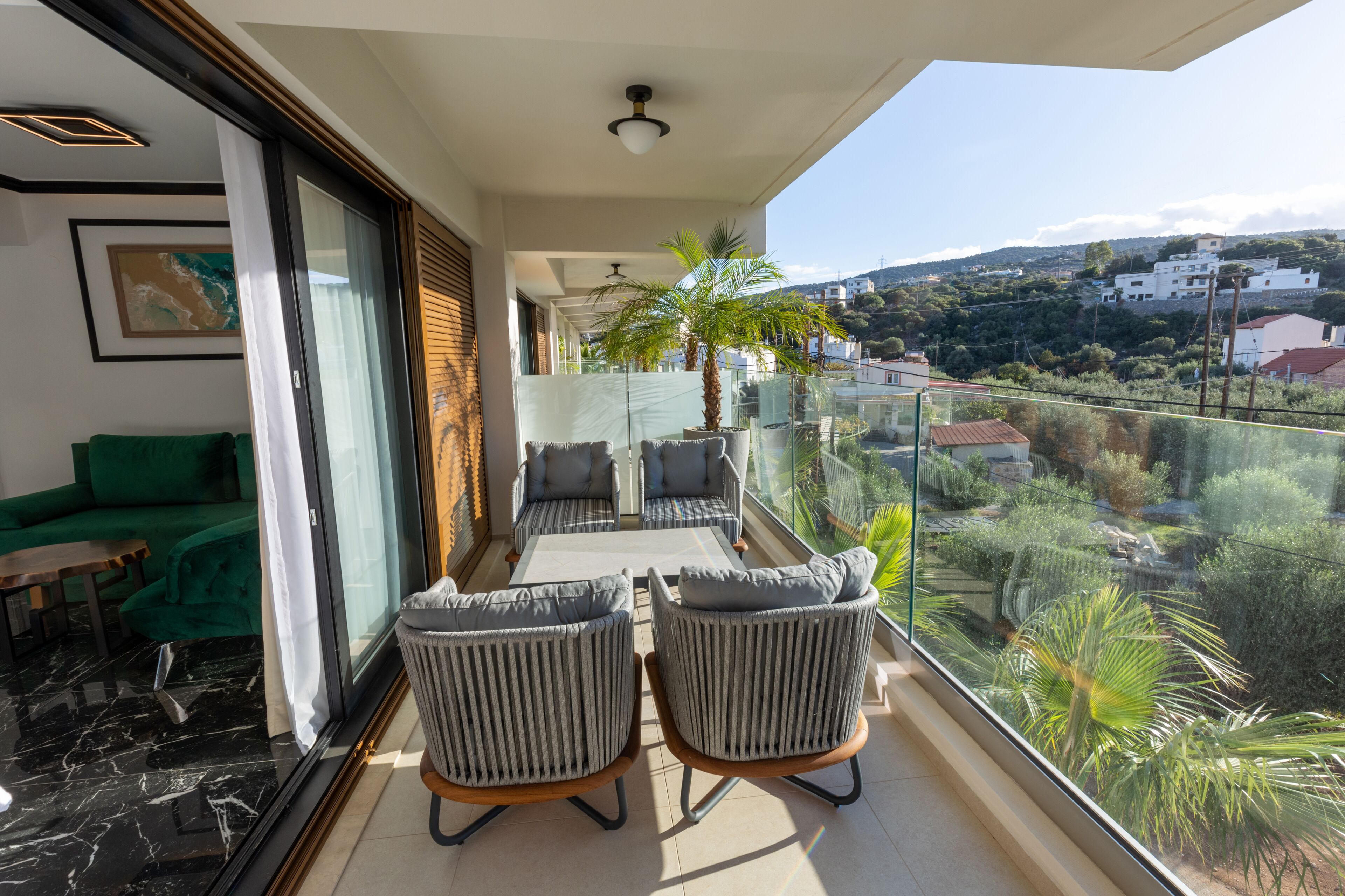 Een balkon met glazen hekwerk, een eettafel met stoelen, een groene bank en uitzicht op een heuvel met gebouwen.