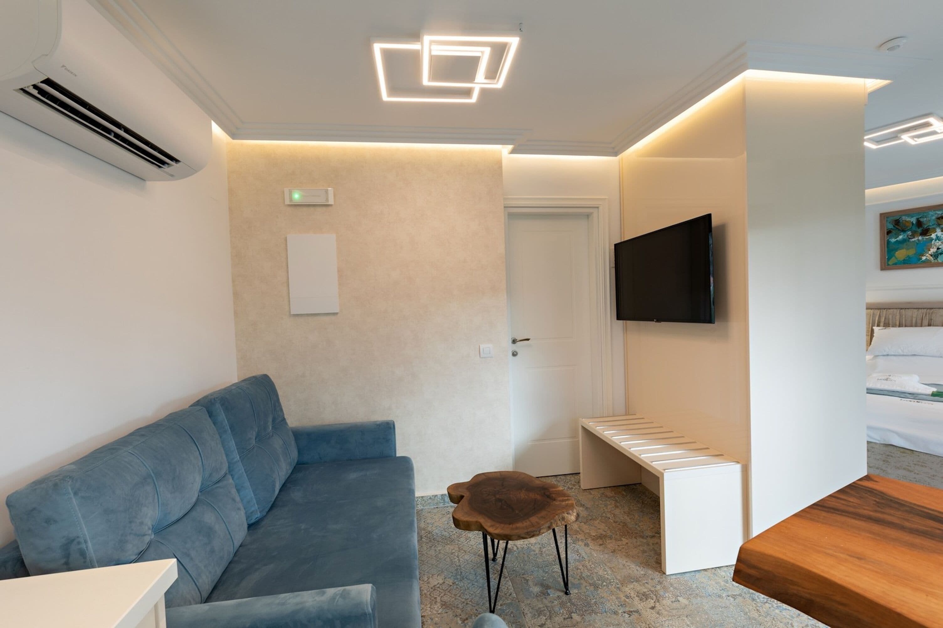 Una habitación de hotel moderna con un sofá azul, una mesita de madera, un banco blanco, un televisor de pantalla plana montado en la pared y una lámpara de techo.