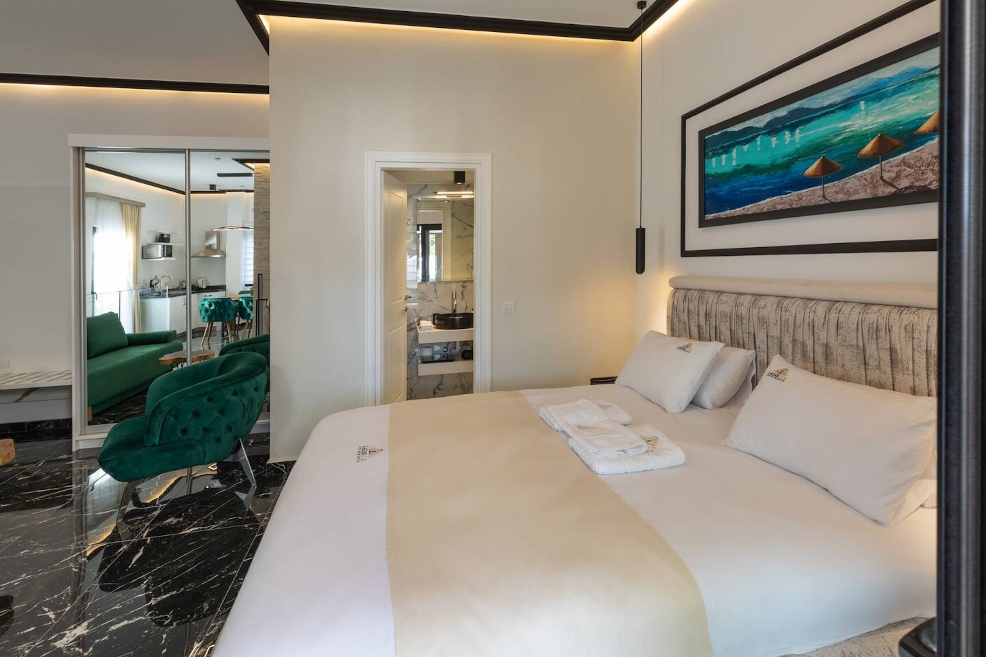 A modern hotel room with a large bed, a green armchair, a painting of a beach scene, and a view into the kitchen area.