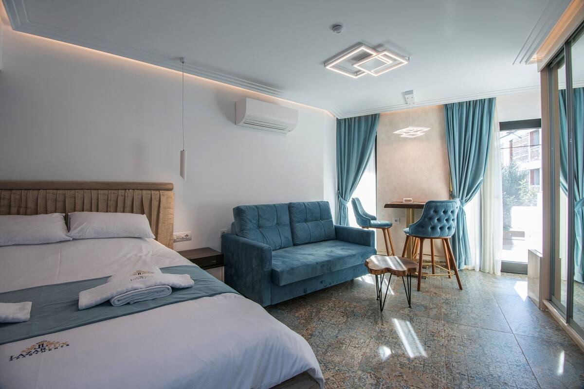 Une chambre d’hôtel avec un lit, un canapé bleu, une petite table avec des chaises et une grande fenêtre avec des rideaux.