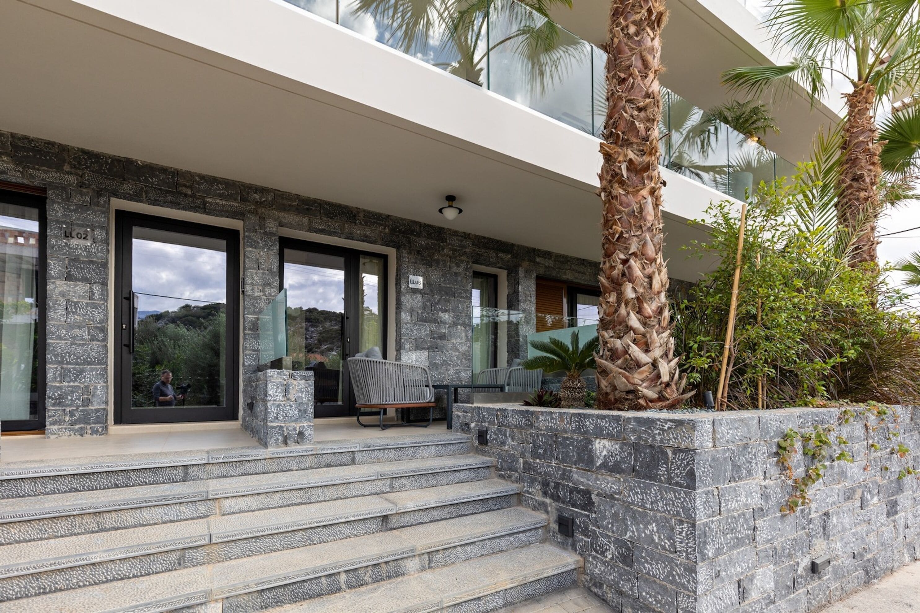 A modern building with stone steps leading to a glass door, outdoor seating, and palm trees.