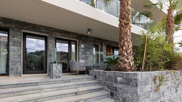 A modern building with stone steps leading to a glass door, outdoor seating, and palm trees.