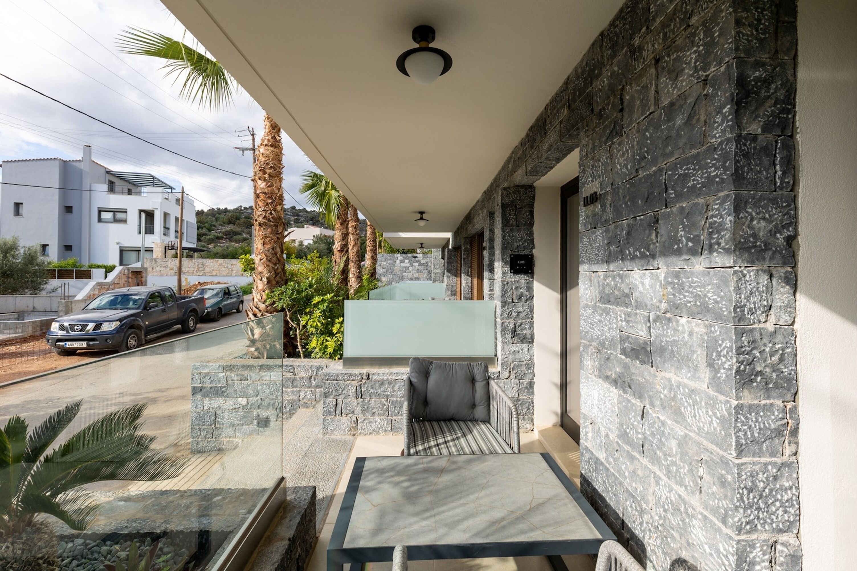 Un balcón moderno al aire libre con paredes de piedra, un ventanal de vidrio y vista a una calle con coches estacionados.