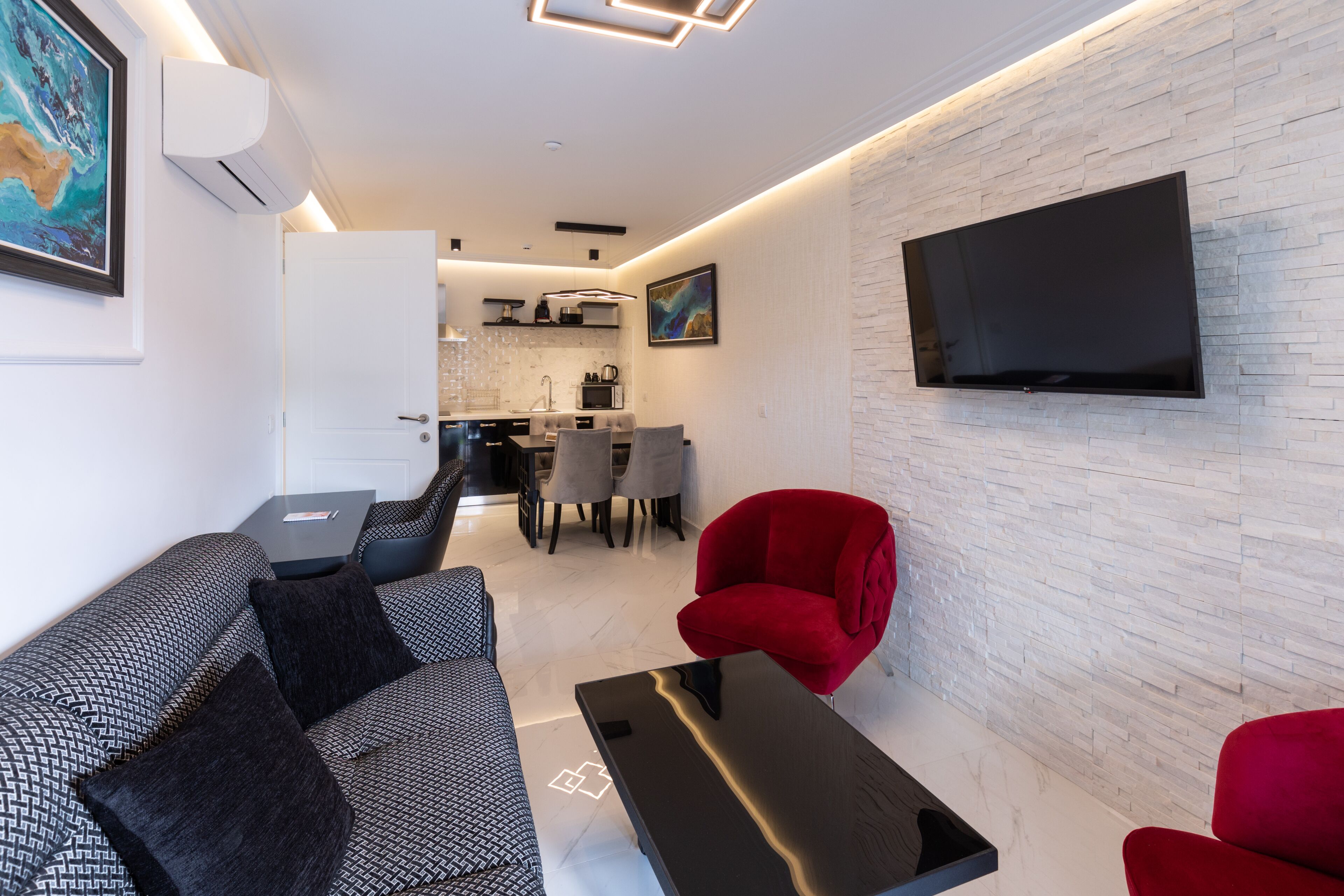 Uma sala de estar moderna com um sofá, uma poltrona vermelha e uma mesa de centro. Uma televisão de tela plana está montada na parede, e há uma área de cozinha ao fundo.