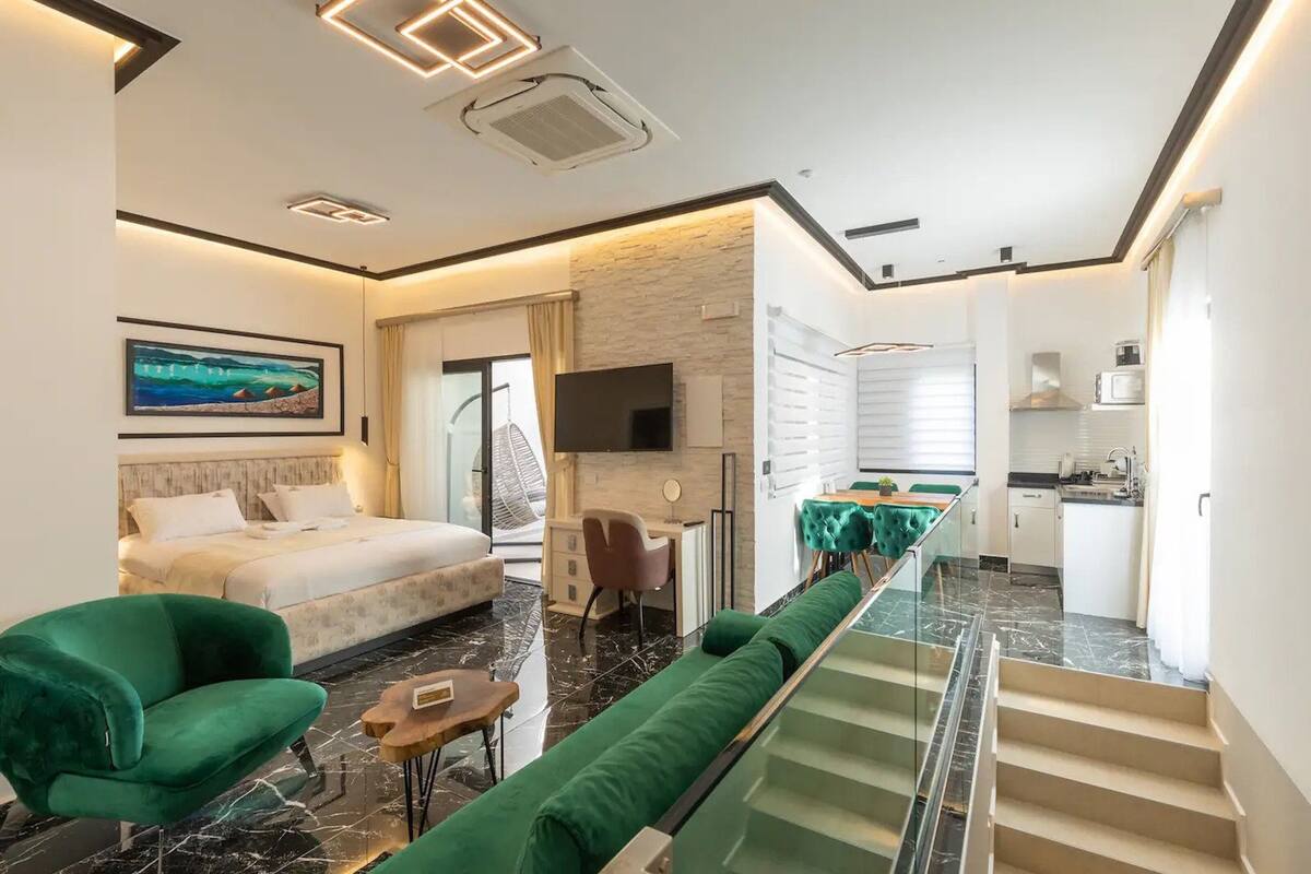 Une chambre d’hôtel moderne équipée d’un lit, de fauteuils verts, d’un canapé, d’un bureau et d’un coin cuisine.