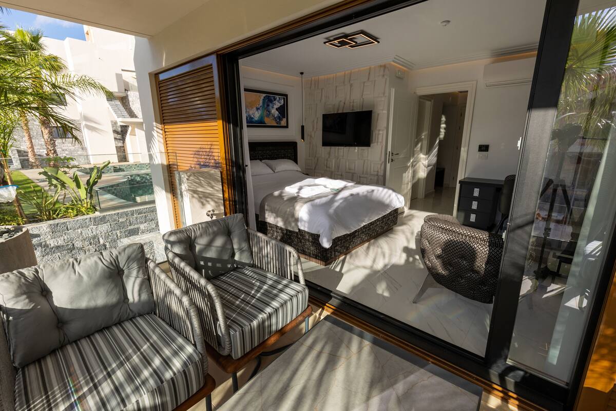 Une chambre moderne avec un lit, une télévision et une porte vitrée donnant sur un balcon équipé d’un canapé et d’un fauteuil.
