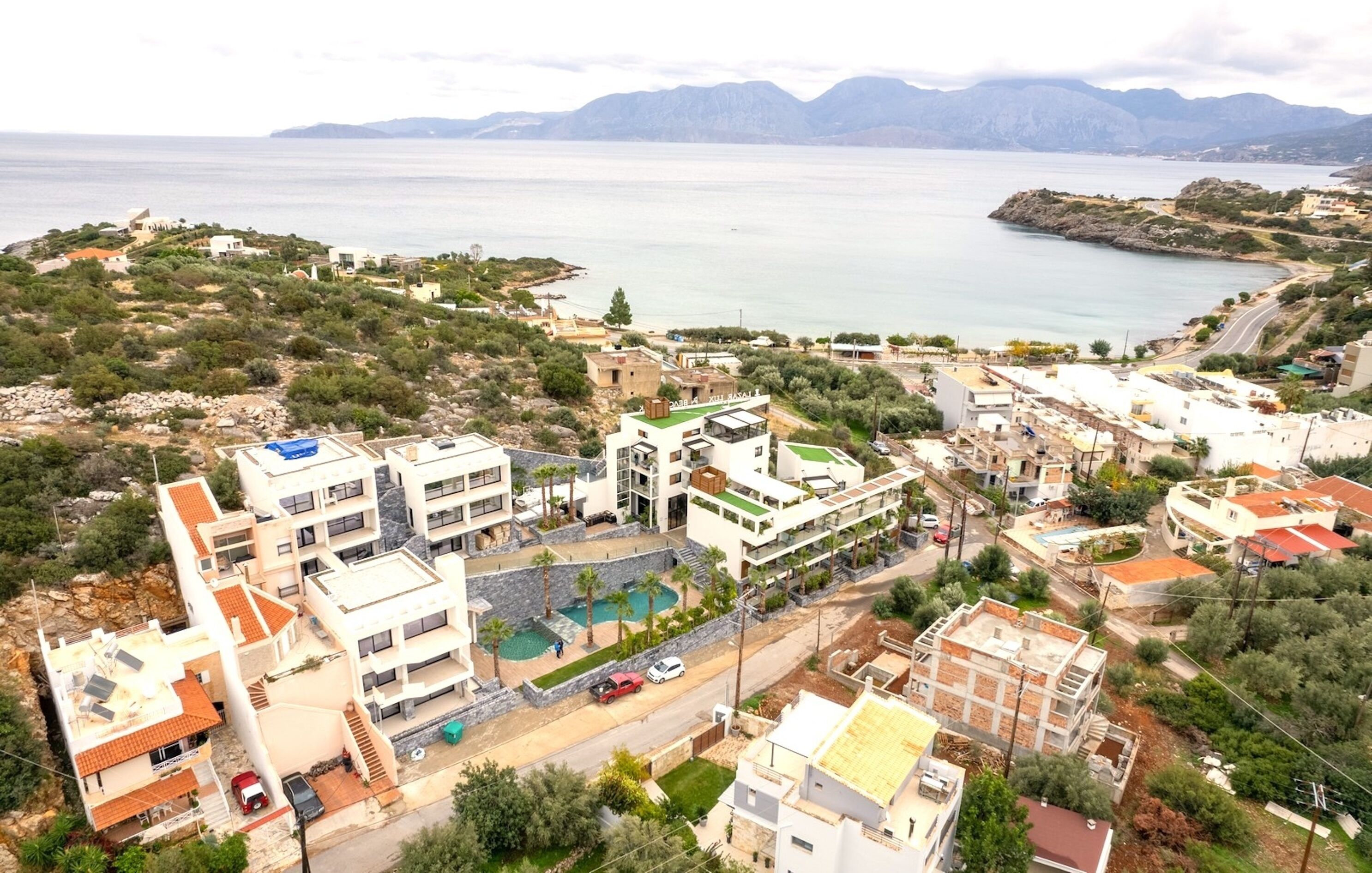 Vista aérea de un pueblo costero con edificios modernos, una piscina y vistas al mar y a las montañas al fondo.