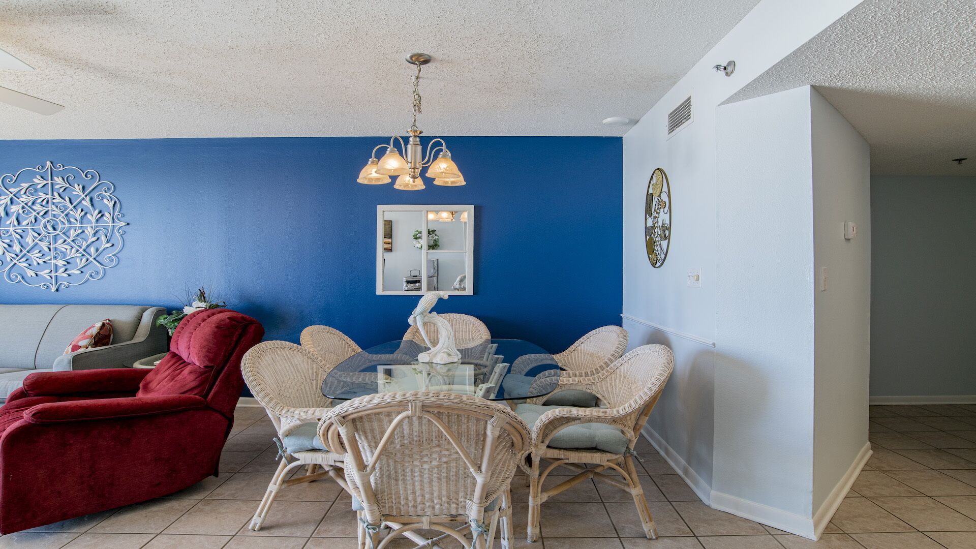Ein Wohnzimmer mit blauer Akzentwand, einem roten Sofa, Korbstühlen, einem Glastisch und einem dekorativen Spiegel.