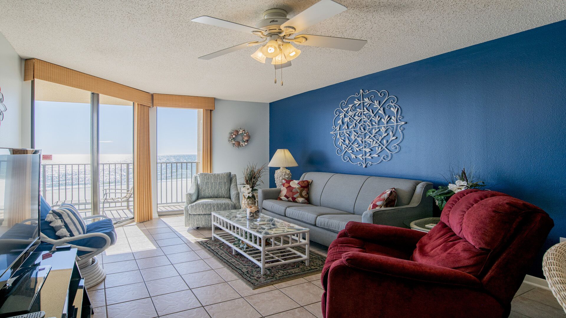 A living room with a blue accent wall, a ceiling fan, a sofa, a red armchair, and a view of the ocean through large windows.