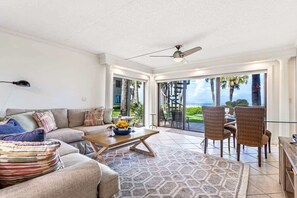 A spacious living room with a sofa, coffee table, and chairs, a ceiling fan, and a large sliding glass door leading to a balcony with a view.