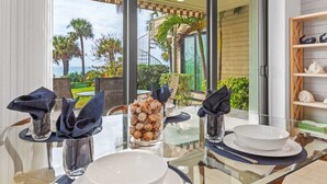 Una mesa de comedor dispuesta con platos blancos, vasos y servilletas, con vista a palmeras y un edificio.