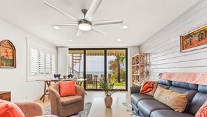 Una sala de estar con ventilador de techo, un sofá, una mesita de centro y vistas al exterior a través de un ventanal.