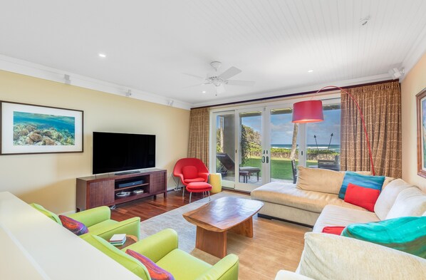 A modern living room with a flat-screen TV, green and white sofas, a wooden coffee table, and a red chair.