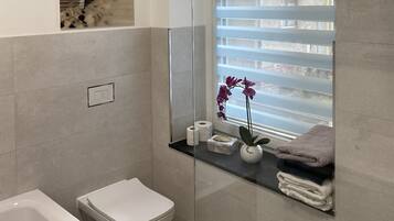 Un baño moderno con cabina de ducha de vidrio, inodoro, bidé y repisa con artículos de aseo.