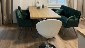 Moderni ruokailutila, jossa on puinen pöytä, kaksi vihreää tuolia, yksi valkoinen tuoli ja seinällä oleva maalaus, jossa on kuvattuna avoin ovi.