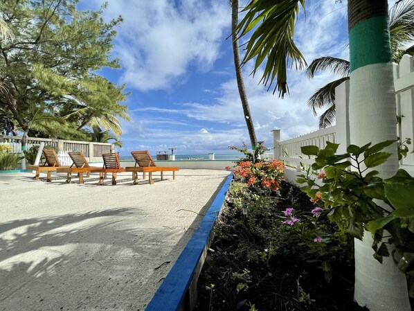 A beachside lounge with wooden chairs and a table, surrounded by plants and a white fence.