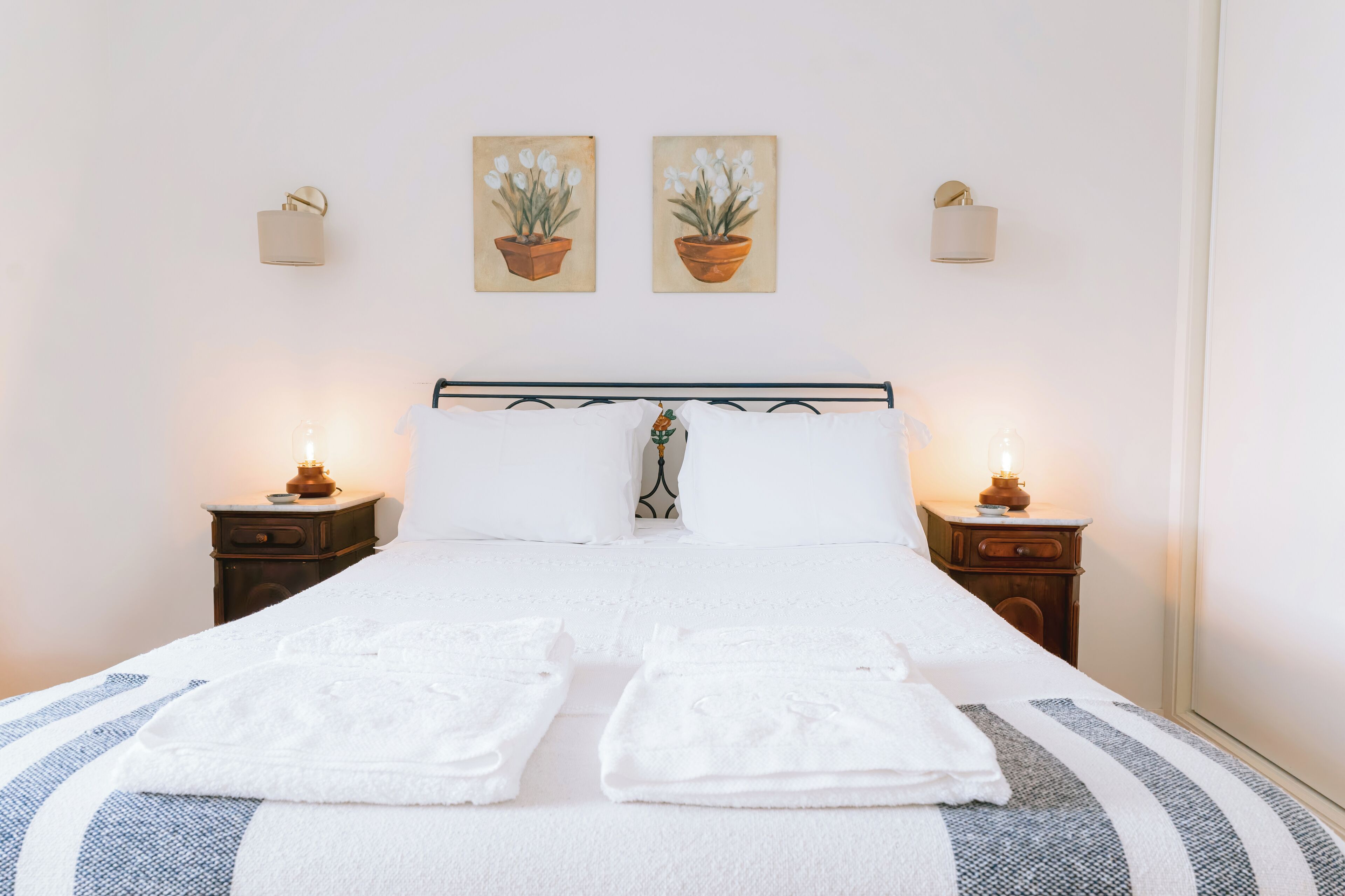 A neatly made bed with white linens and towels, flanked by two bedside tables with lamps, in a room with plain walls and two potted plants on the wall.