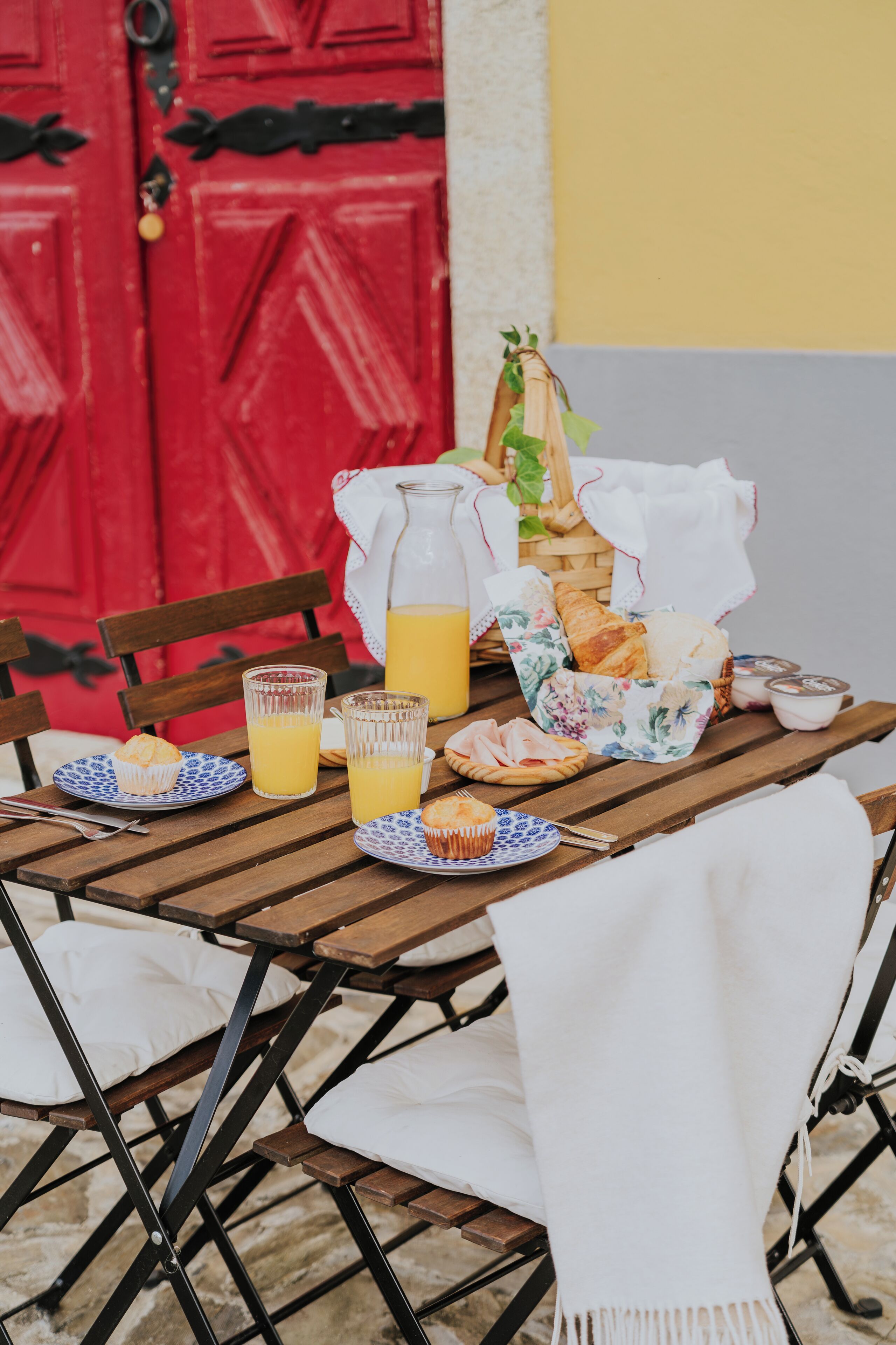 A wooden table set with breakfast items including croissants, muffins, and juice, with a red door and yellow wall in the background.