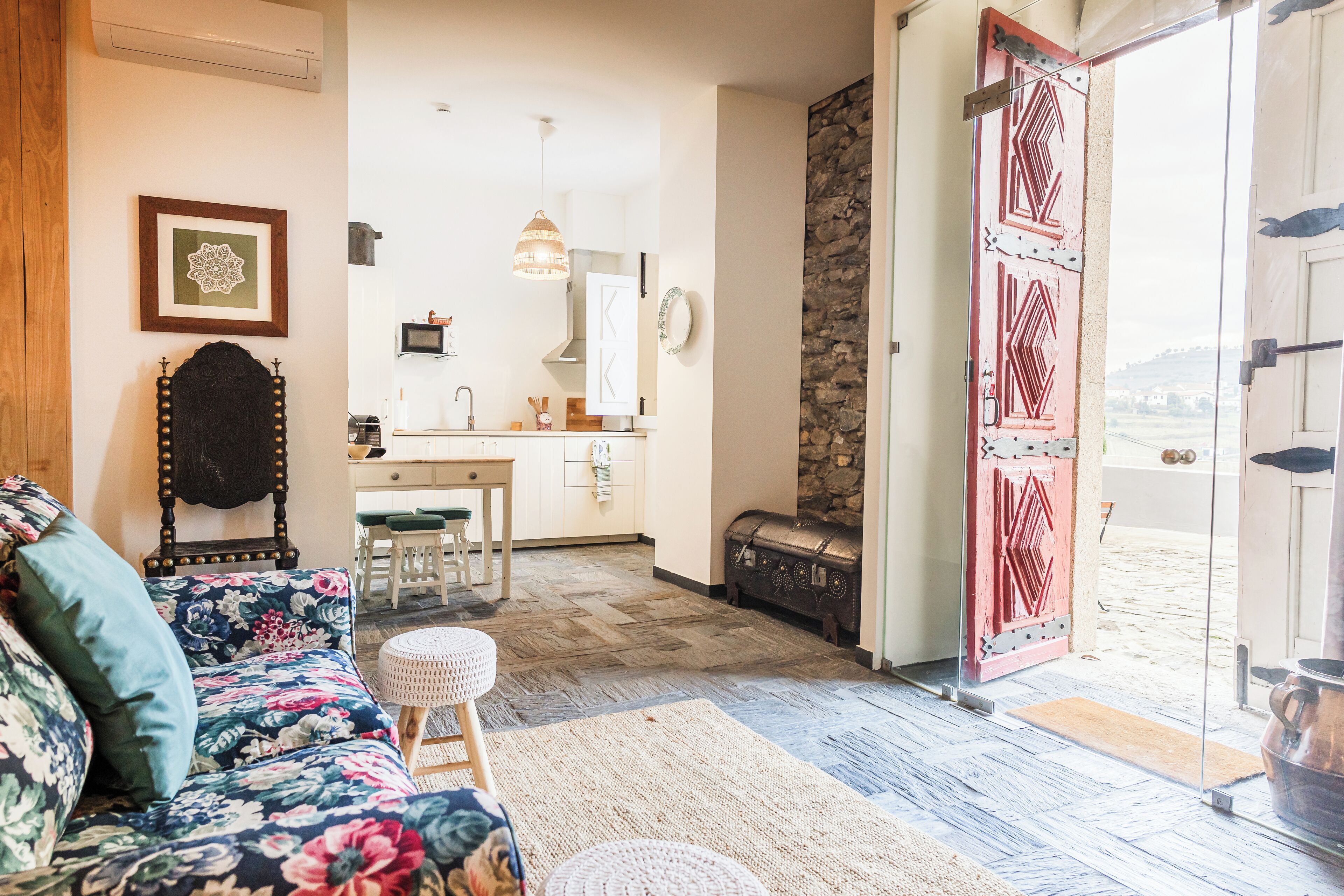 Ein gemütliches Wohnzimmer mit einem blumigen Sofa, einem Holzstuhl und einem kleinen runden Tisch. Im Hintergrund ist eine Küchenzeile mit einem Esstisch und Stühlen zu sehen.