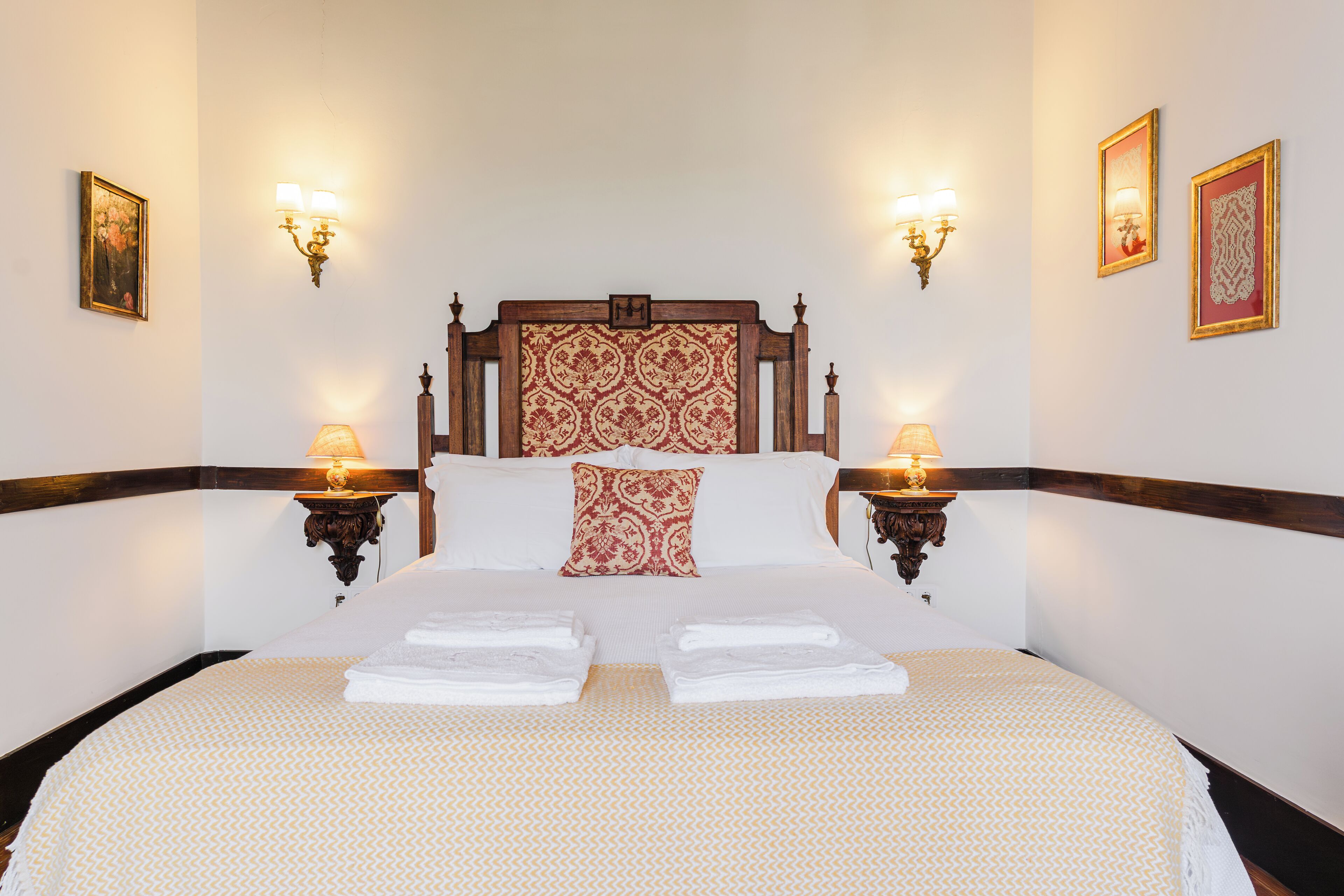 Una cama bien hecha con sábanas blancas y dos almohadas, flanqueada por mesitas de noche con lámparas, en una habitación con cabecera de madera y obras de arte enmarcadas en las paredes.