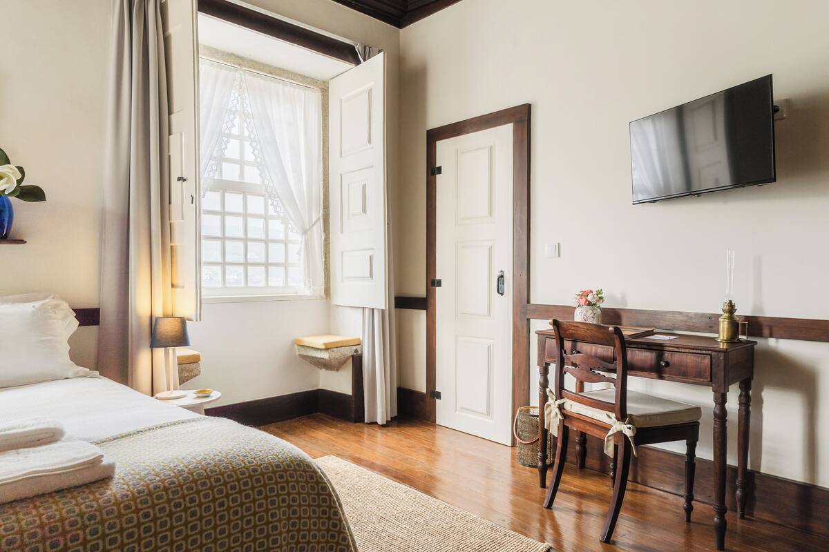 Quarto de hotel com cama, escrivaninha com cadeira, televisão e janela com cortinas.