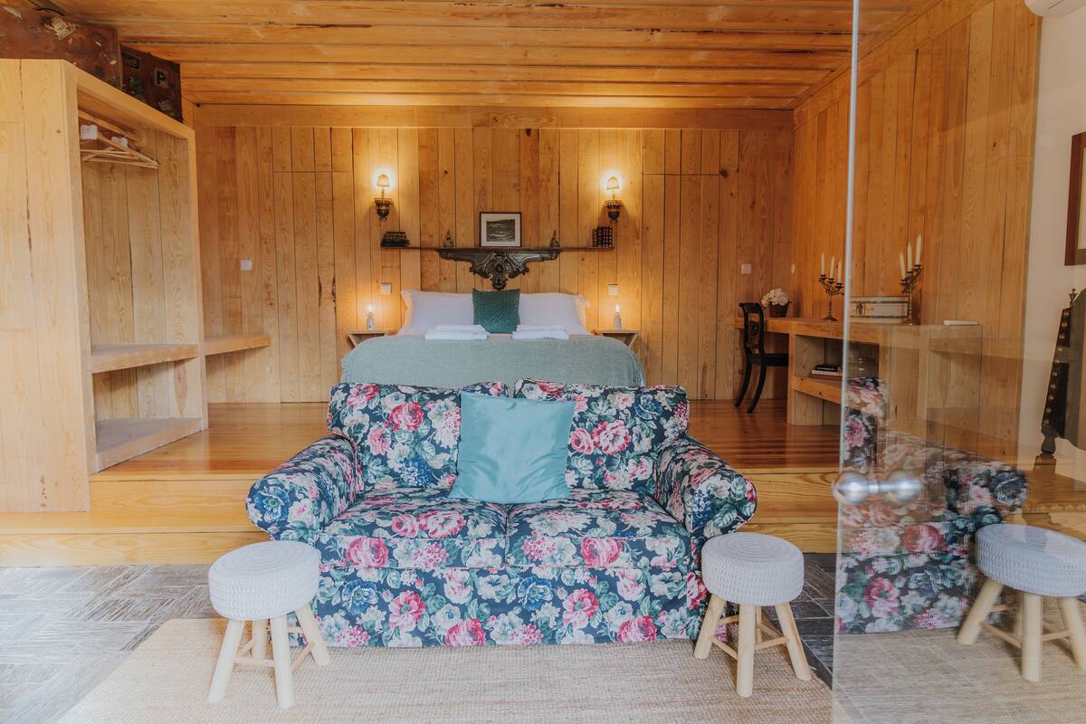 Quarto com painéis de madeira, uma cama, um sofá floral, duas mesinhas de cabeceira e uma pequena mesa com velas.