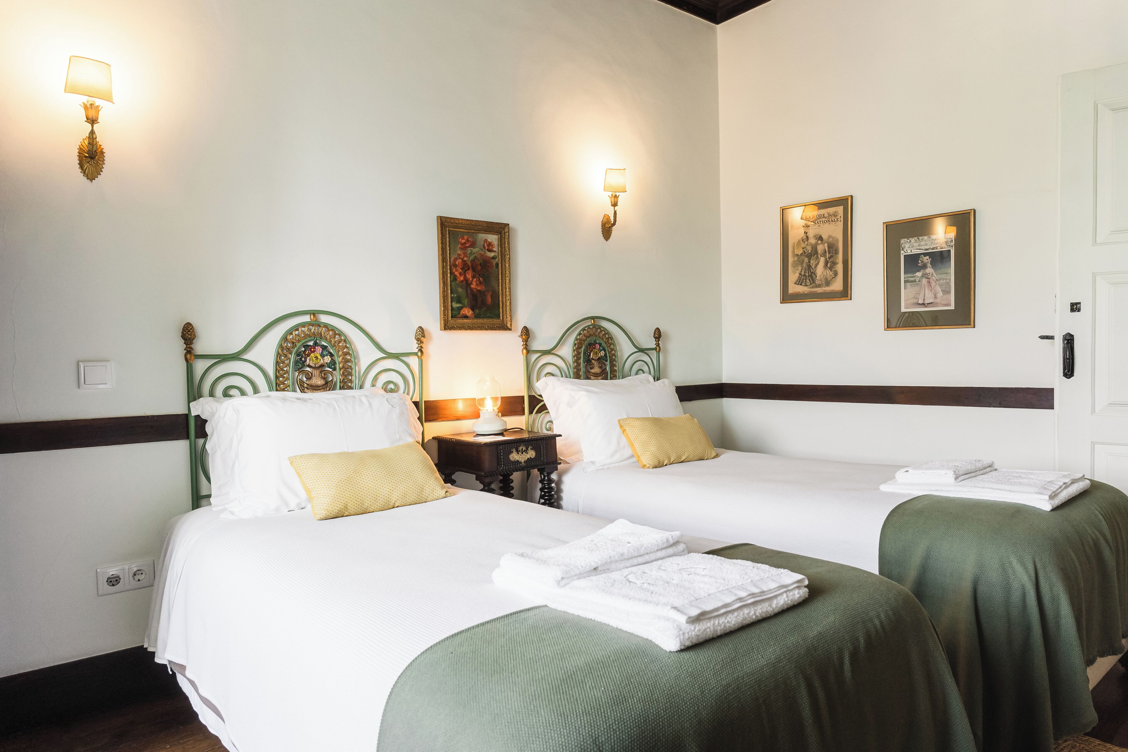 A hotel room with two beds, green bedspreads, white linens, and wall-mounted lamps.
