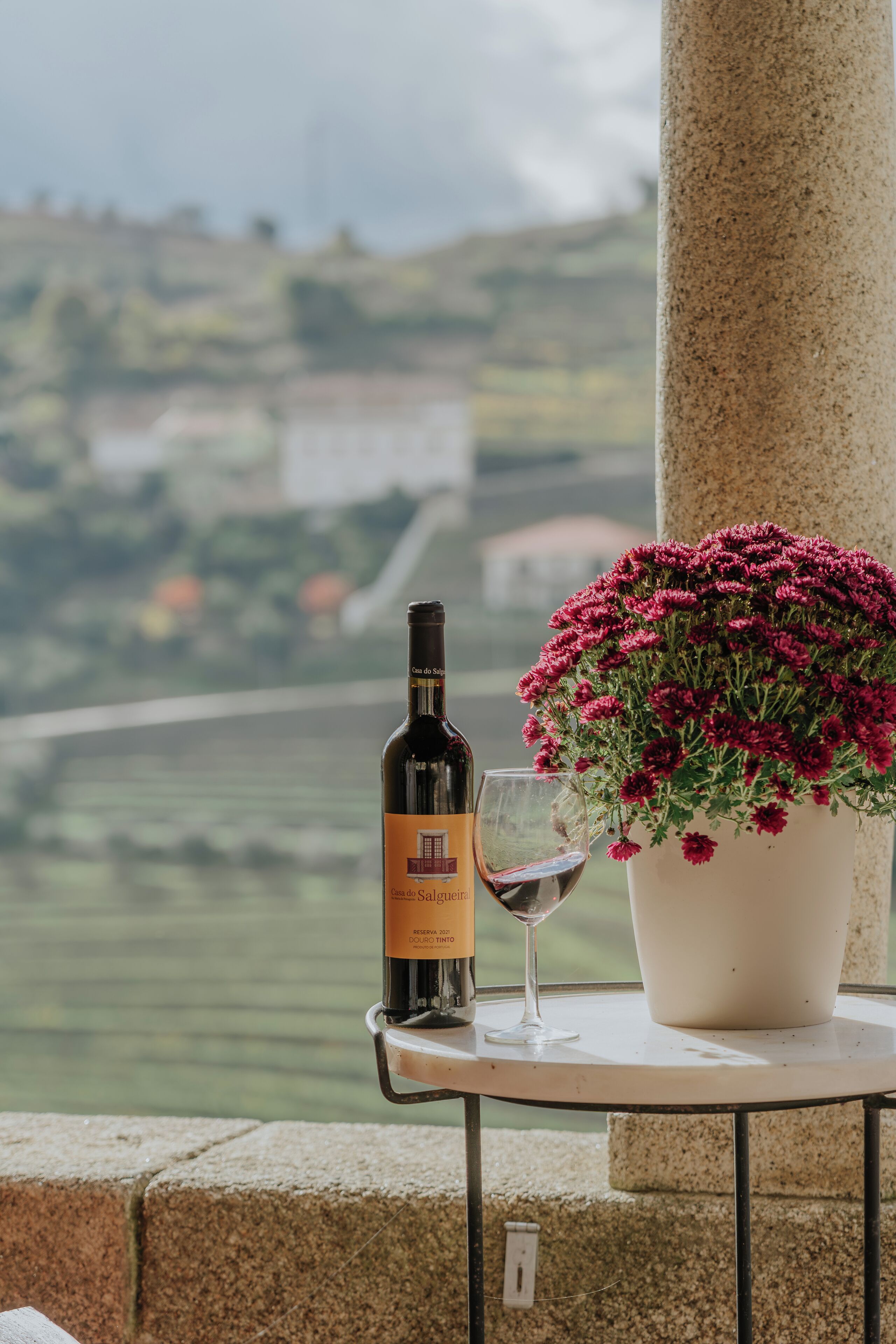 A bottle of wine and a glass on a table with a view of a vineyard.