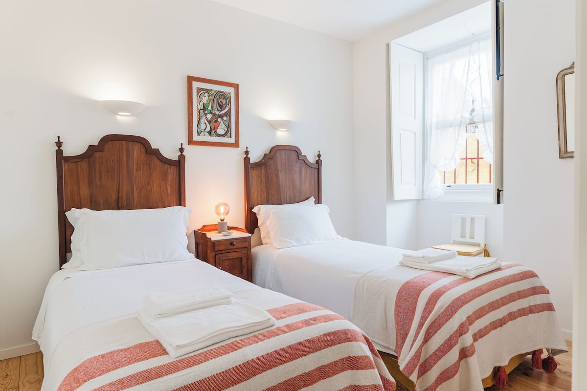 Quarto de hotel com duas camas, cada uma com cabeceira de madeira, roupa de cama branca e manta listrada em vermelho e branco. Há uma mesa de cabeceira com uma luminária entre as camas e uma janela com vista para um edifício.