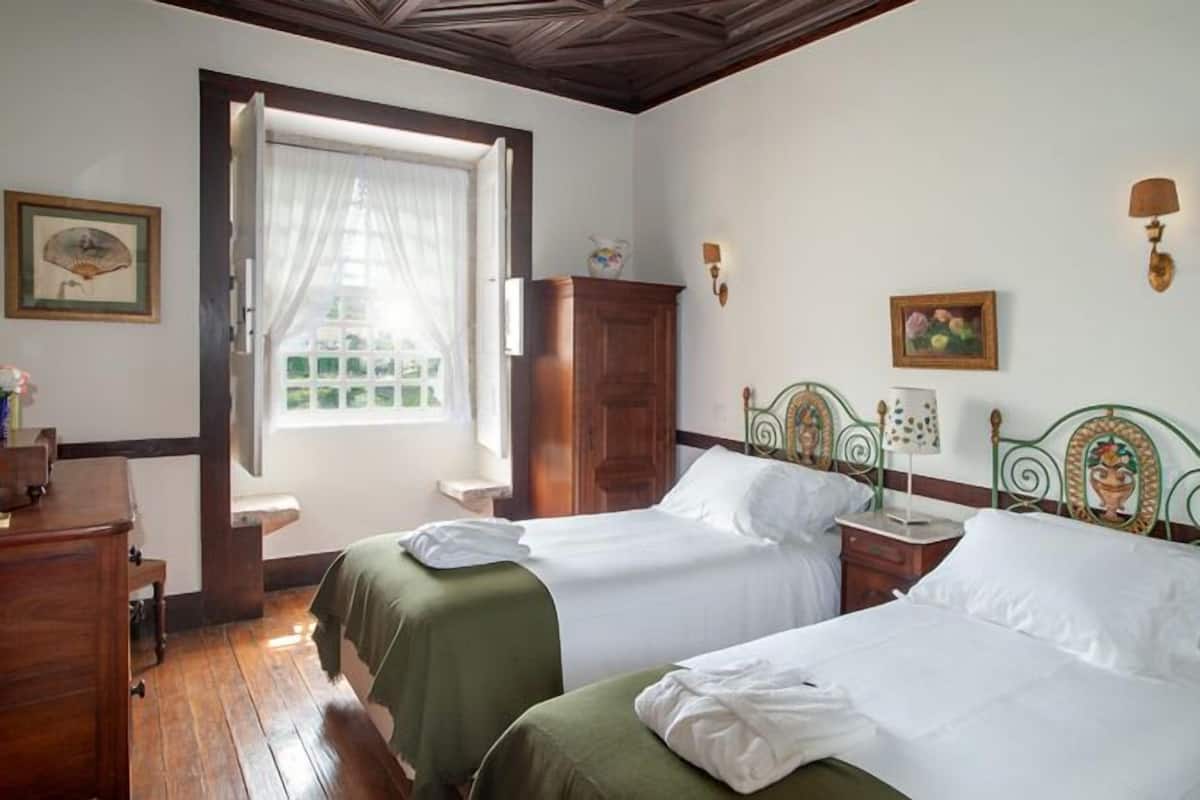 Quarto com duas camas, guarda-roupa de madeira, janela com cortinas, luminárias fixadas na parede e quadros emoldurados.