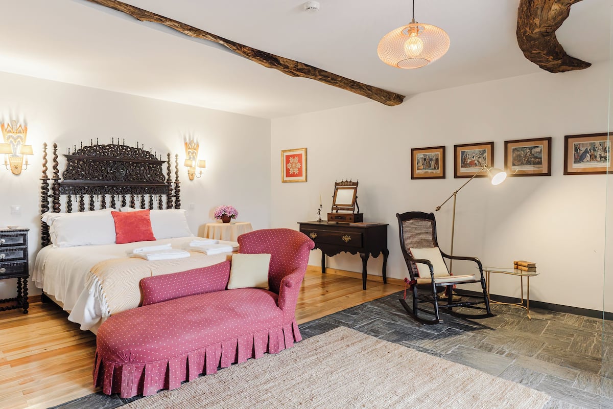 Quarto com uma cama grande, um banquinho vermelho com bolinhas, cabeceira de madeira e espelho fixado na parede.