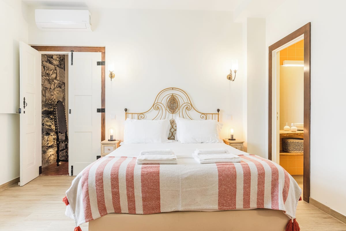 Cama bem arrumada com manta listrada, parede de pedra atrás da porta e banheiro visível por uma porta aberta.