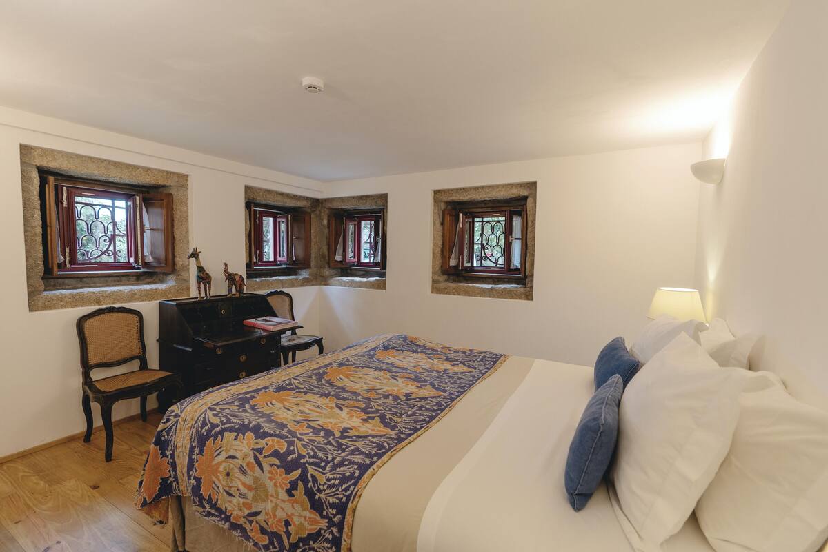 Ein Schlafzimmer mit einem Bett, einem Schminktisch, einem Stuhl und drei mit Vorhängen versehenen Fenstern.