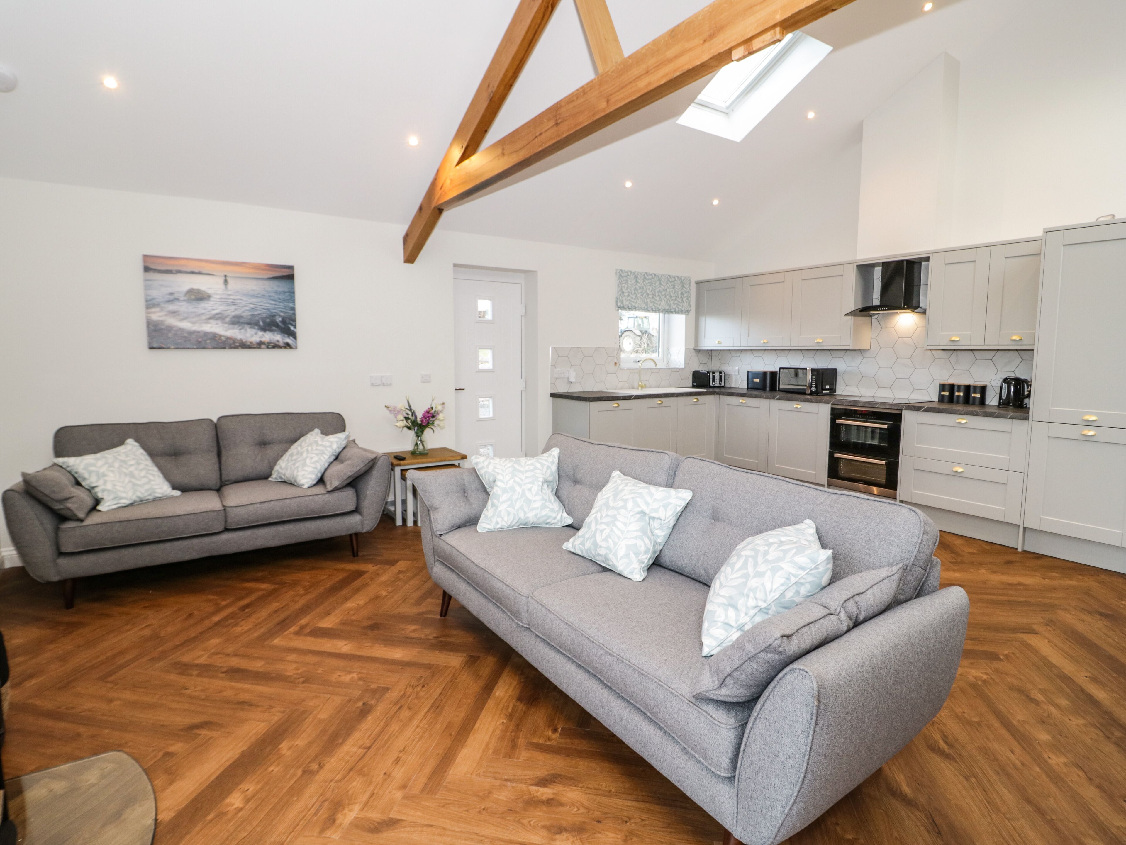 A modern living room with two grey sofas, a wooden floor, and a kitchen with white cabinets and a black stove.