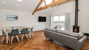Un salon moderne avec un plancher en bois, un canapé gris, une table à manger avec des chaises, un téléviseur fixé au mur et un poêle à bois.