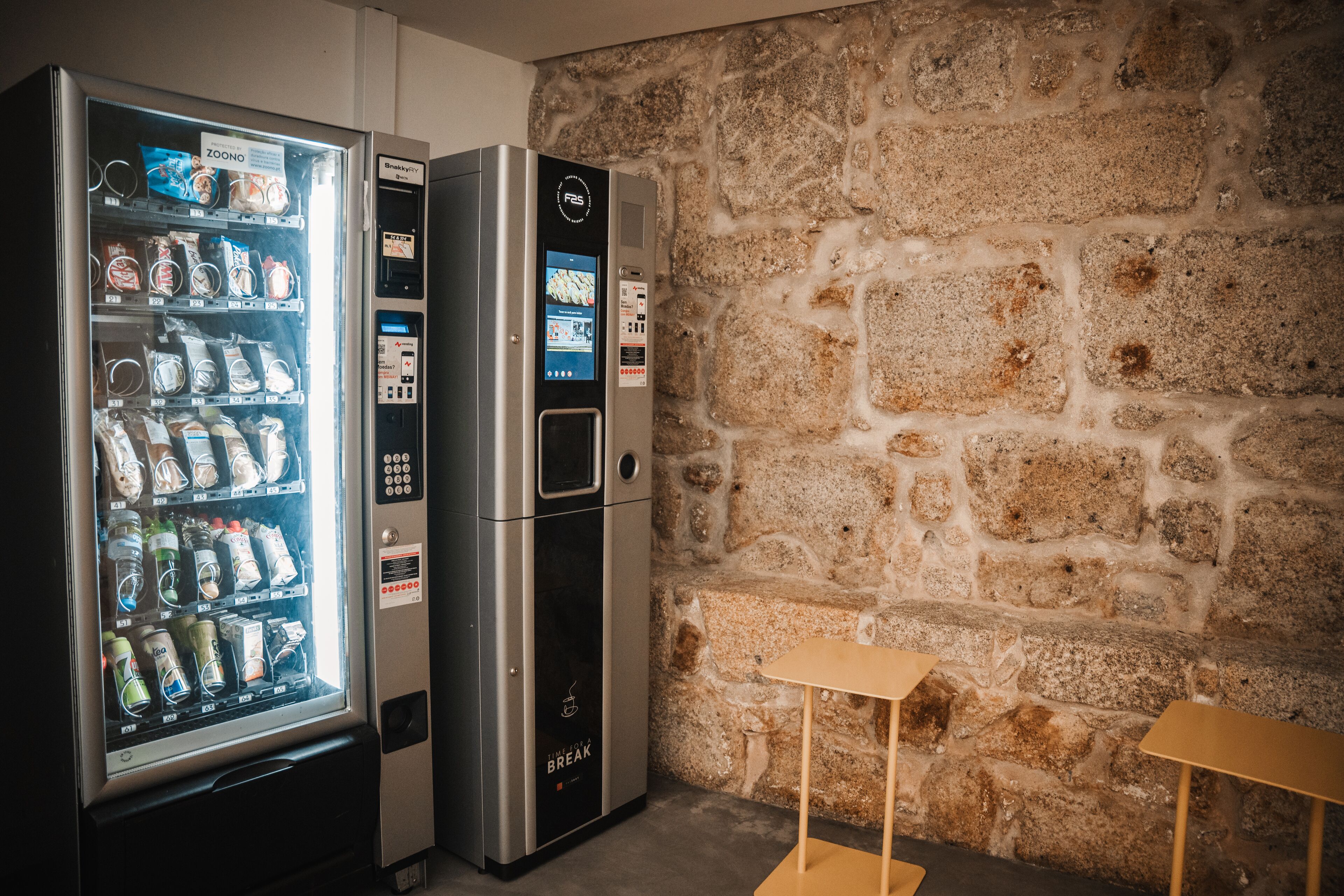 A vending machine with various snacks and drinks, a digital payment screen, and a stone wall in the background.