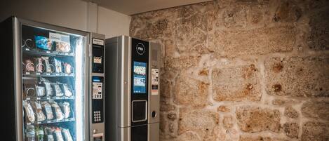 Egy automatás árusító, amelyben különféle rágcsálnivalók és italok kaphatók, egy digitális fizetési képernyő, és a háttérben egy kőfal látható.