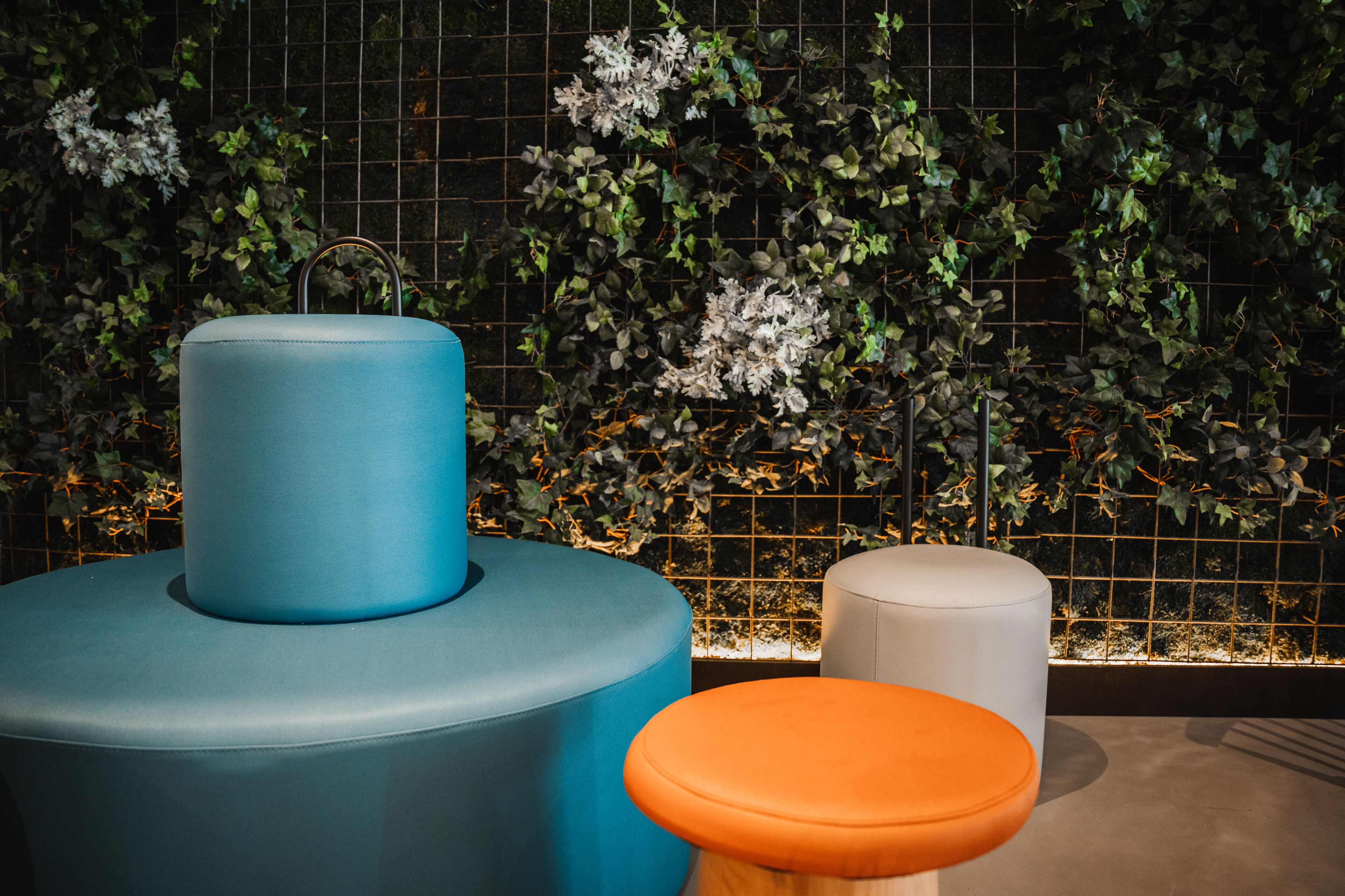 Un área de estar moderna con un reposapiés azul y naranja, un taburete blanco y una pared decorativa con plantas y flores.