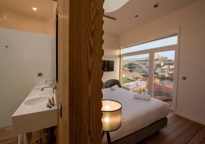 A modern bedroom with a large bed, a wooden door, a bathroom with a sink, and a city view through large windows. - The Central House Porto Ribeira - Hostel (Porto)