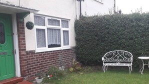 Una casa con puerta verde, ventana blanca con cortinas de encaje, banco de metal blanco y un seto.