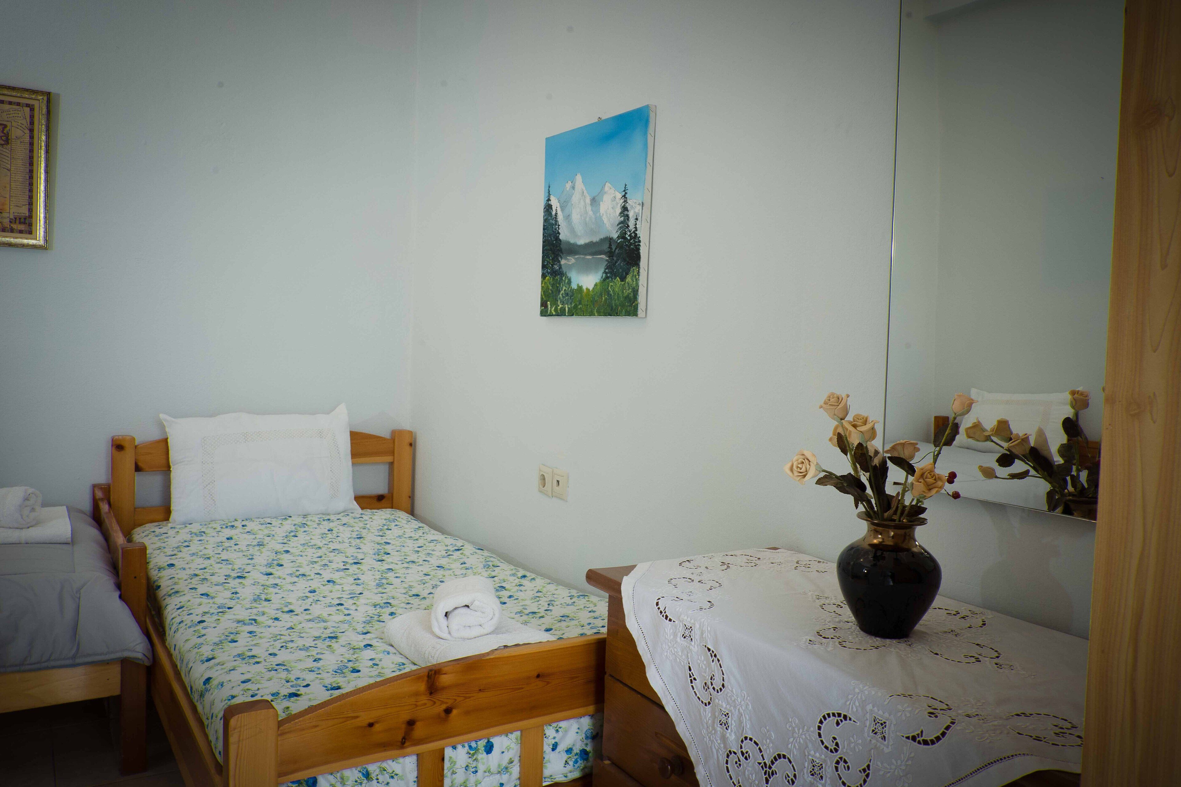 Une chambre à coucher avec un lit en bois, une petite table sur laquelle est posé un vase de fleurs, et un tableau représentant un paysage de montagne accroché au mur.