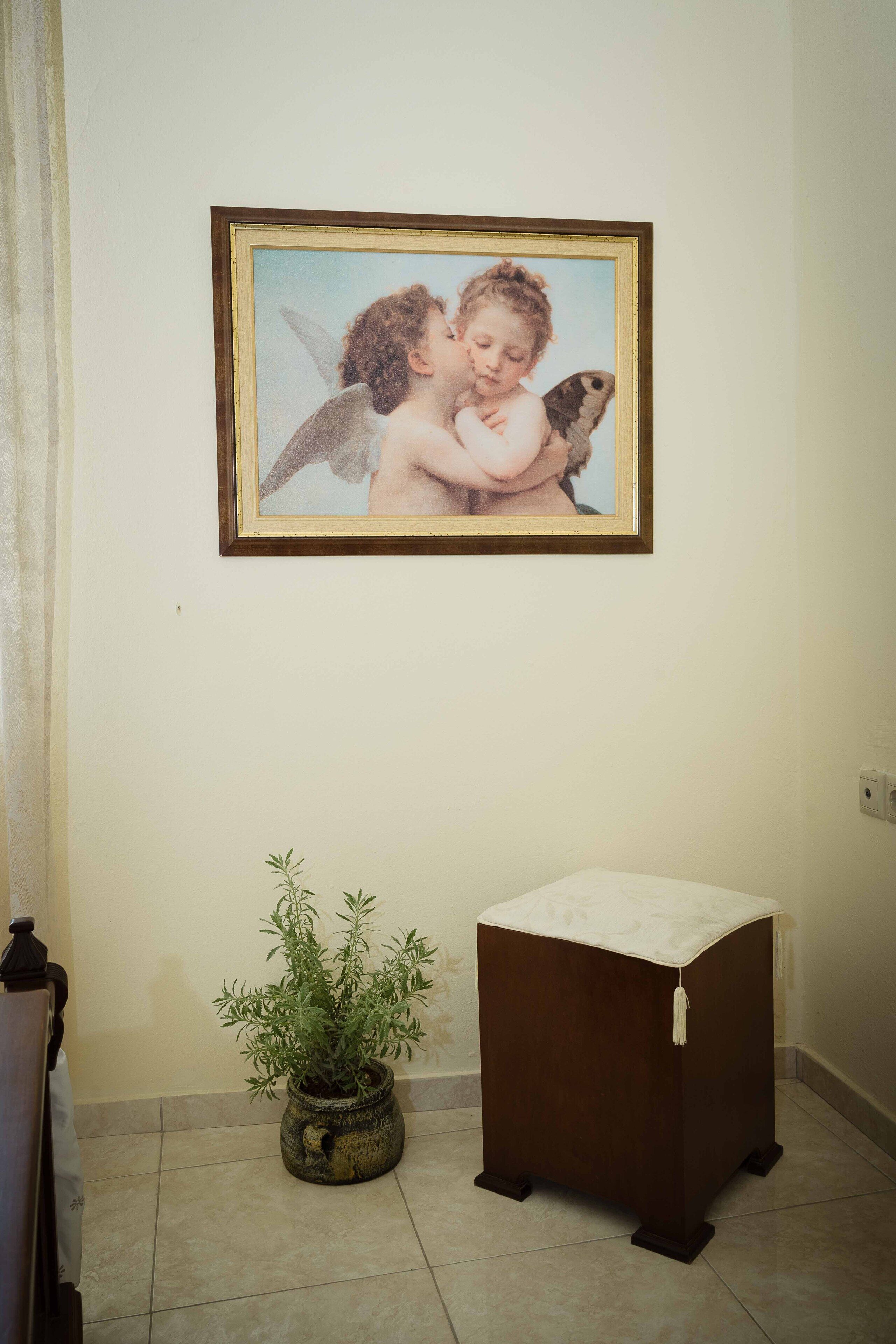 A framed artwork of cherubs on a wall, a small potted plant, and a bedside table with a white cloth.