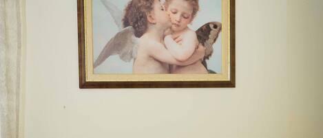 A framed artwork of cherubs on a wall, a small potted plant, and a bedside table with a white cloth.