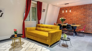 Una sala de estar con un sofá amarillo, una mesa de comedor con sillas, una cortina roja y una pared de ladrillo.