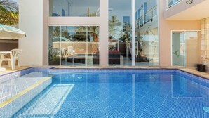 A modern pool area with clear glass walls, a tiled pool, and a white outdoor seating area. - Beachfront House (SP)