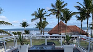 A terrace with a black table and chairs, overlooking a beach with palm trees and a red-roofed building. - Beachfront House (SP)