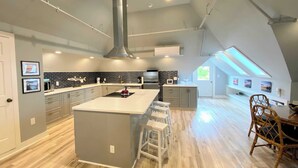 A modern kitchen with a large island, a dining area with wicker chairs, and a view of the outdoors through a skylight. - Channel View Residence at the Dygert House (Clayton)
