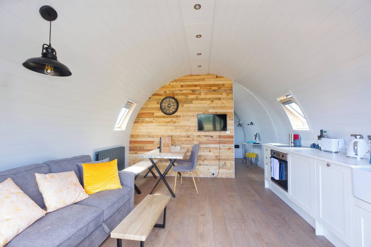 A compact kitchen with a wooden wall, a dining table, and a sofa with cushions.