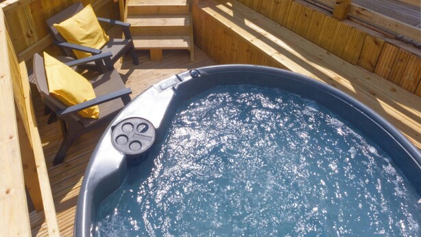 Una terraza de madera con un jacuzzi, una escalera de madera y una habitación acristalada con vistas al paisaje.